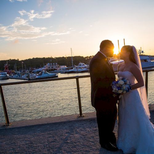 A bride and groom stand by a waterfront at sunset, holding hands and gazing at each other, with boats docked in the background.