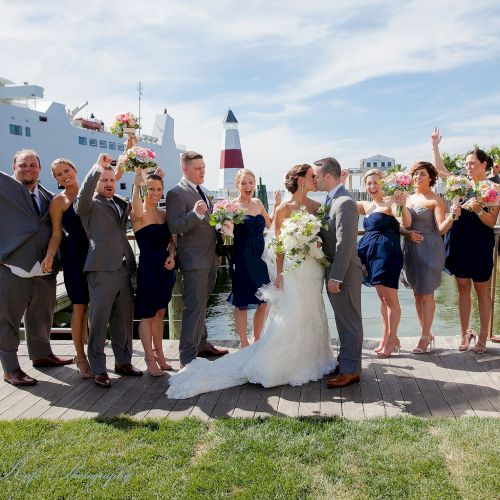 A wedding party celebrates by the waterfront, with the bride and groom kissing in the center and a large ship in the background.