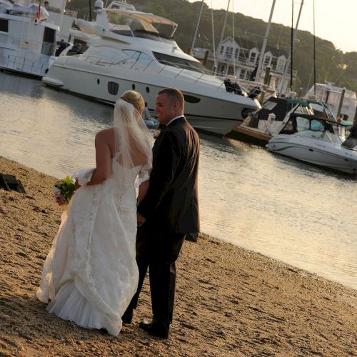 A couple in wedding attire stands on a sandy shore, overlooking boats docked in a marina under a setting sun.