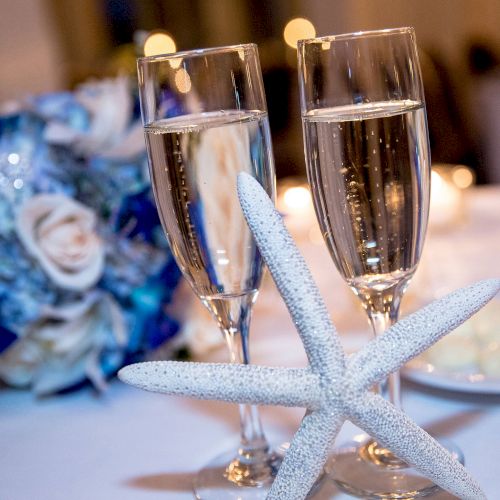 Two champagne flutes with a decorative starfish in front, a blue and white bouquet in the background, and blurred lights in the distance.