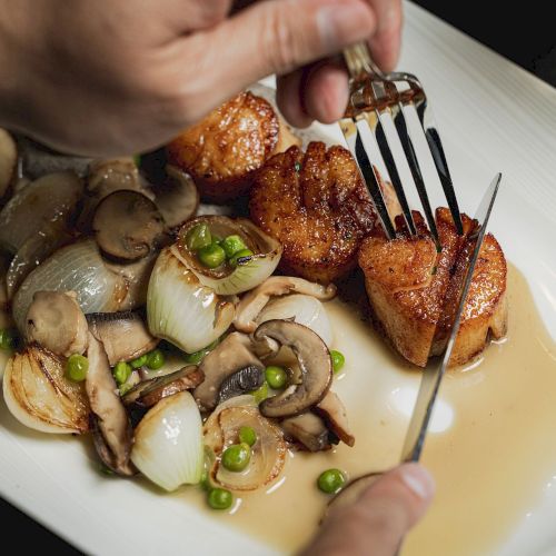 A person is cutting and eating scallops on a plate with sautéed mushrooms, onions, and peas, all in a light sauce.