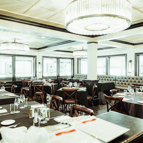 A spacious, elegantly lit dining area with multiple tables set with glasses and plates, featuring modern chandeliers and large windows.