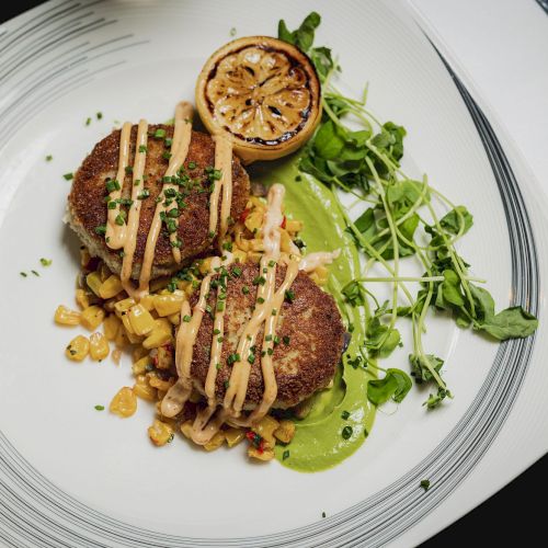 The image shows a gourmet dish consisting of two patties topped with sauce, sitting on a bed of corn, and garnished with greens and a grilled lemon half.