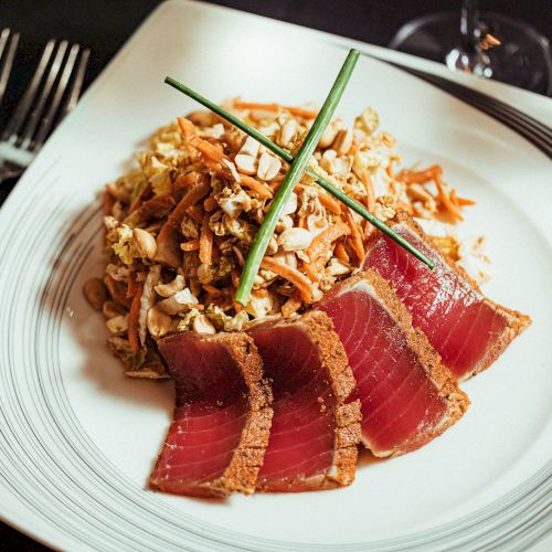 The image shows a dish with slices of seared tuna, garnished with chives and accompanied by a mixed salad on a white plate with a fork and knife beside it.