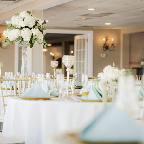 The image shows an elegant, white-themed banquet or wedding reception setup with round tables, floral centerpieces, and neatly arranged tableware.