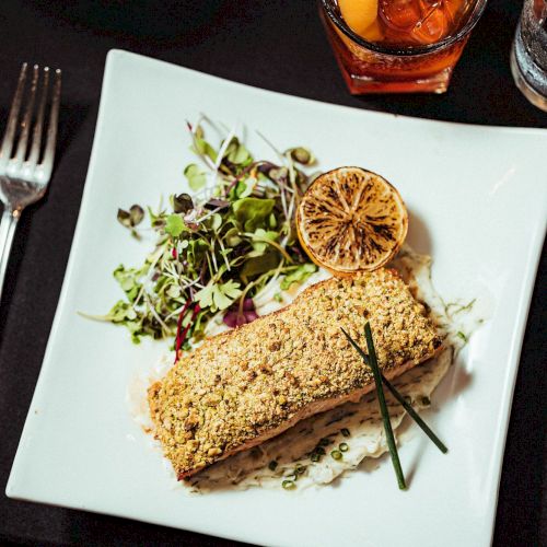 A plated meal includes a piece of crusted fish, a lemon slice, microgreens, with forks and a knife at the sides, and a drink in the background.