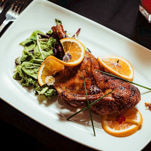 A beautifully plated roasted chicken garnished with orange slices, served alongside a fresh green salad with utensils beside the plate.