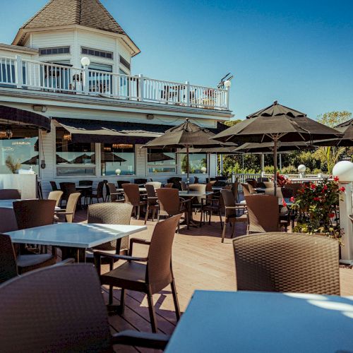 Outdoor restaurant patio with wicker chairs, tables, and black umbrellas, set next to a building with a second-story deck. Clear blue sky.