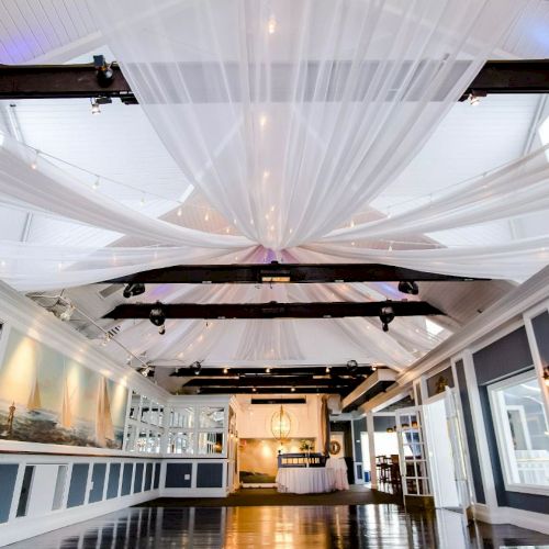 The image shows an elegantly decorated event space with white drapes on the ceiling, cushioned seating along the walls, and a polished wooden floor.