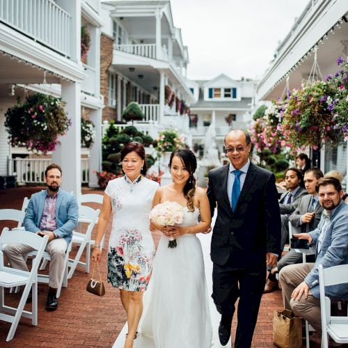 A bride in a white dress, holding a bouquet, walks down an outdoor aisle with two people, surrounded by seated guests and flower decorations.