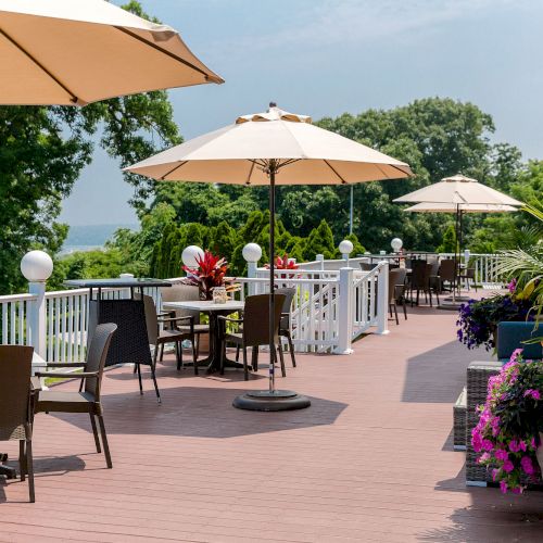 An outdoor patio with tables and chairs, large umbrellas for shade, surrounded by greenery and flowers on a sunny day.