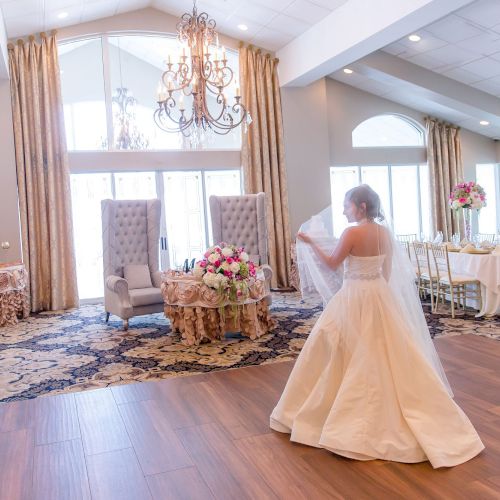 A bride in a white dress stands alone in a spacious, elegantly decorated room with chandeliers, flowers, and large windows, looking at two chairs.