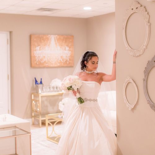 A bride in a white dress stands in a room adorned with empty picture frames and elegant decor, holding a bouquet of flowers.