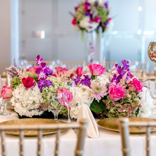 The image shows an elegant event table setup with a flower arrangement of pink, white, and purple flowers, surrounded by glasses and decorative gold items.