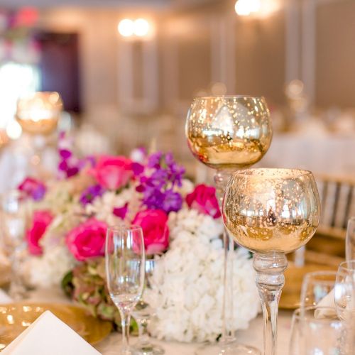 The image shows an elegantly set table featuring gold goblets, champagne flutes, and a vibrant floral centerpiece in a softly-lit, formal setting.