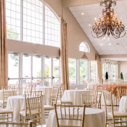 A spacious, elegant banquet hall with large windows, round tables covered with white linens, gold chairs, and a grand chandelier hanging from the ceiling.