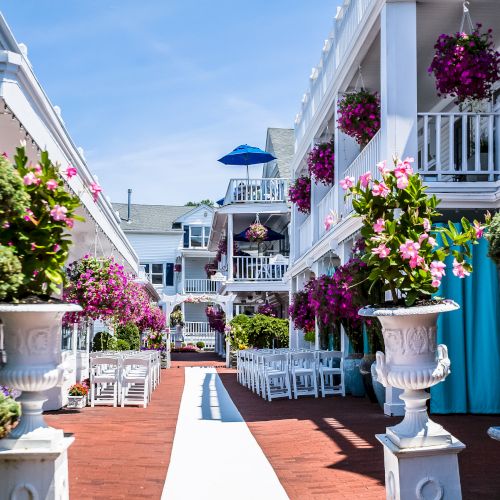 A courtyard with white buildings, balconies adorned with pink flowers, trimmed greenery, tables, and chairs lined up along a red brick walkway.