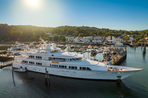 The image shows a large white yacht docked at a marina with other boats and a scenic town in the background under a clear sky.