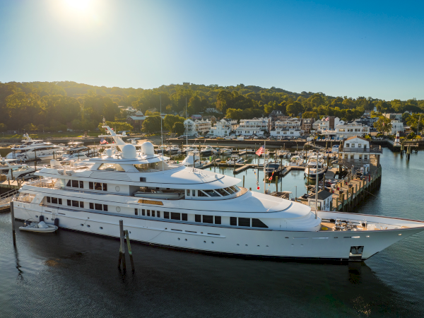 The image shows a large white yacht docked at a marina with other boats and a scenic town in the background under a clear sky.